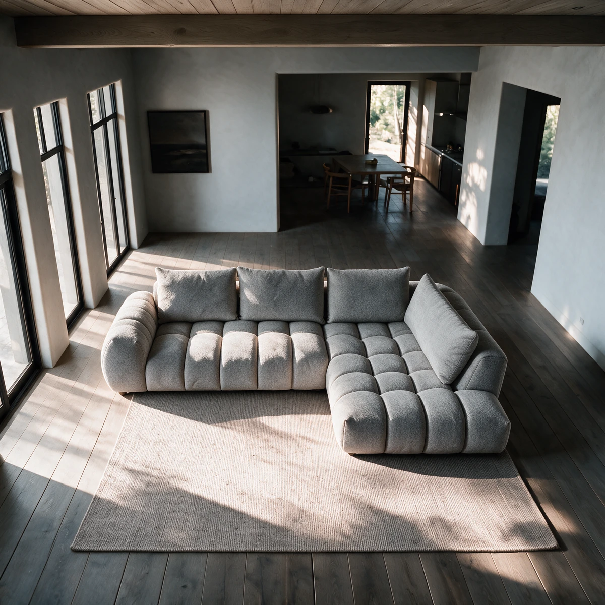 A grey corner sofa with soft, sculpted seat blocks and a chaise, seen from above on a large rug in a sunlit room with tall windows